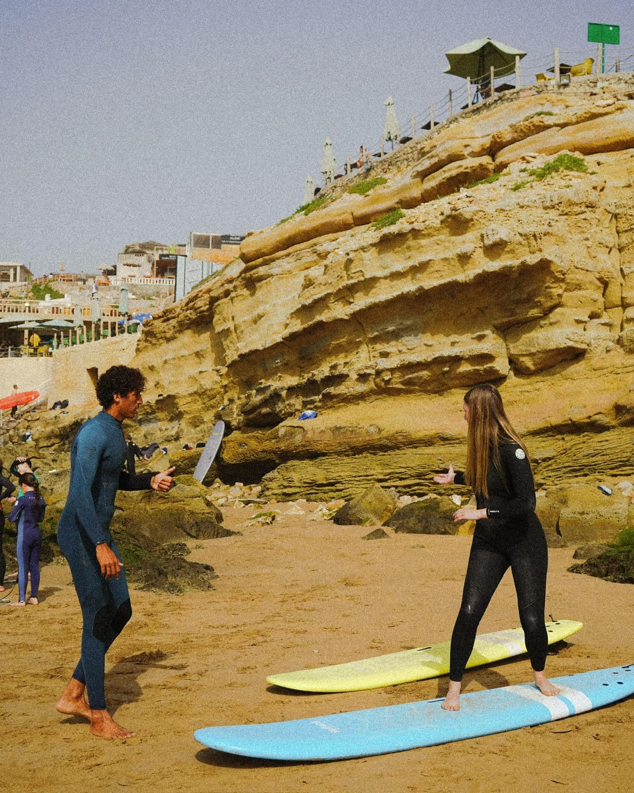 A coach demonstrating the pop-up stance to a surfer standing on a longboard on the sand, cliffs behind